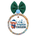 I'm freakin freezin lettering with a bundled-up Highland cow in a blue winter hat and scarf, surrounded by snowflakes.