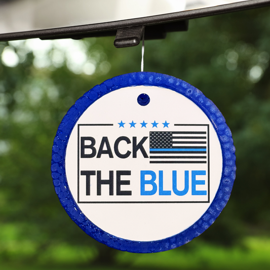 Round 'Back the Blue' air freshener hanging from a car's rearview mirror with a blurred green background.