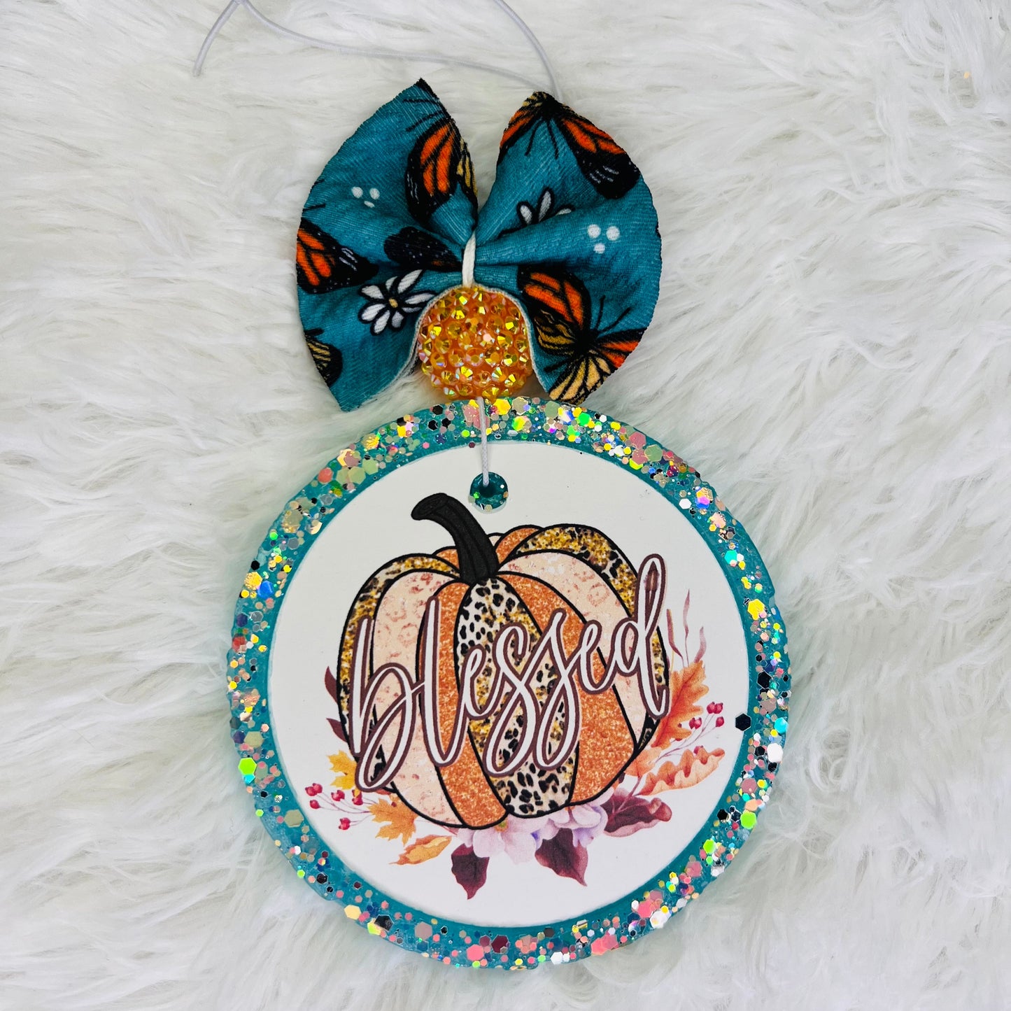 A decorative air freshener featuring a pumpkin design with a butterfly, leaves, and the word 'Blessed' written on it, adorned with multicolored beads.
