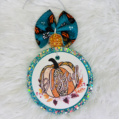 A decorative air freshener featuring a pumpkin design with a butterfly, leaves, and the word 'Blessed' written on it, adorned with multicolored beads.
