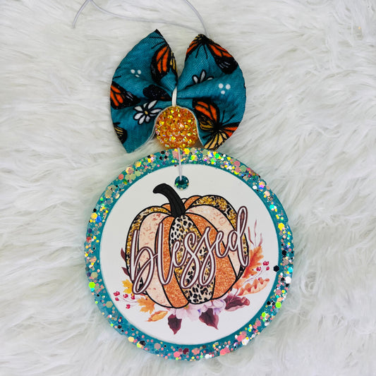 A decorative air freshener featuring a pumpkin design with a butterfly, leaves, and the word 'Blessed' written on it, adorned with multicolored beads.