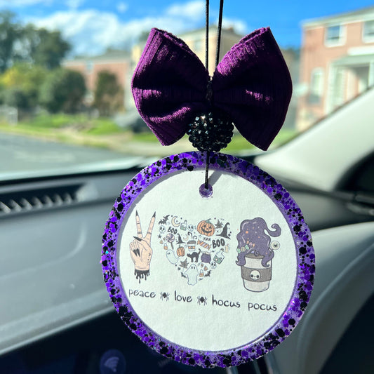 A purple-colored air freshener with a bow and a cartoonish illustration that includes the text 'PEACE LOVE HOCUS POCUS' and various Halloween-themed images like witches and ghosts.