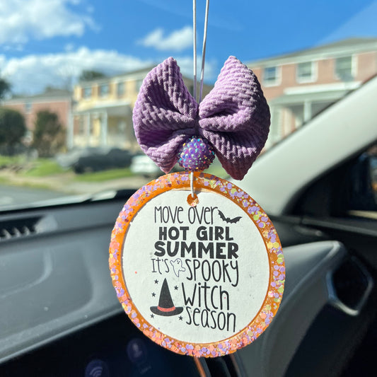 A decorative air freshener with a witch-themed design, featuring a purple bow and orange beads, hanging from a car's rearview mirror.