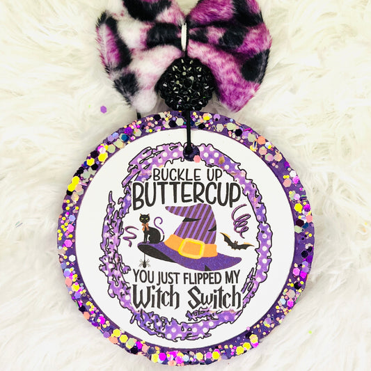 A round air freshener with a witch and buttercup graphic, featuring purple and black colors and text, with a purple bow on top.